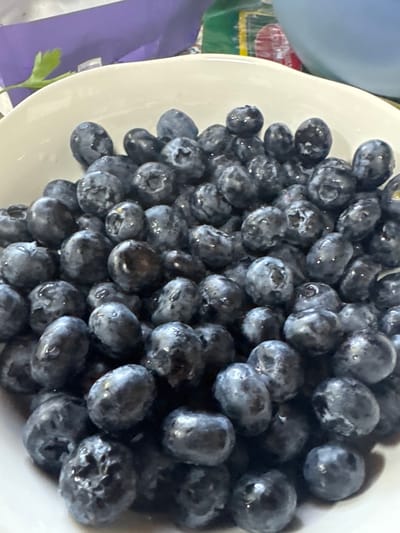 A white bowl is filled with a large quantity of blueberries. The berries are dark blue with a powdery bloom, and some appear to have water droplets on their surfaces. The bowl is sitting on a surface near a blue object, a purple object, and some greenery. There is no indication this photo was taken in Малая Рукавицкая, Russia.