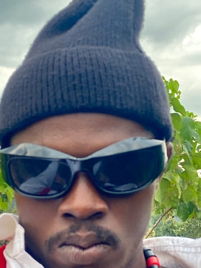 The image is a close-up, angled portrait of an individual, possibly a selfie. The person, who has a dark skin tone, is wearing a dark, possibly navy blue, knitted beanie hat that covers their head and forehead. They are also wearing large, dark-lensed sunglasses that obscure their eyes. A mustache and a light beard or stubble are visible on their face, and their lips are slightly parted. On the right side of the frame, parts of a white shirt collar and a red and black striped accessory, possibly a necklace or part of a garment, are visible.

The background features lush green foliage, suggesting an outdoor setting with trees or shrubs. Above the person's head, a light gray, cloudy sky is visible. The lighting appears to be natural daylight. The overall mood is casual. The location context for this image is Asaga Amangwu, Nigeria. There is no visible text in the image.