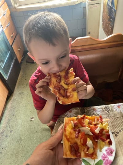 A young boy, with brown hair and fair skin, is seated at a table and eating a slice of pizza. He is wearing a red t-shirt. In the foreground, a hand holds another slice of pizza on a white plate with a floral pattern. The setting appears to be a home interior. The background shows wooden furniture, tiled walls, and a window. The floor is covered with a green rug. The image is taken from a slightly high-angle perspective, looking down at the boy and the table.
