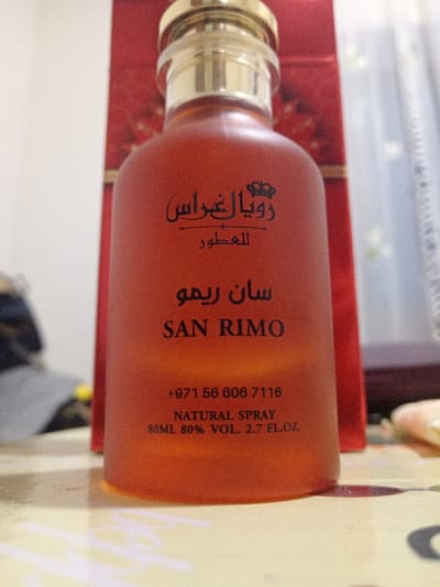 An orange, frosted bottle of "SAN RIMO" perfume is displayed prominently. Above the name is the Arabic text "رويال غراس" and "للعطور". Below "SAN RIMO" is the phone number "+971 56 606 7116" and the words "NATURAL SPRAY" followed by the volume "80ML 80% VOL. 2.7 FL.OZ.". The bottle has a gold cap. A red box is visible in the background.