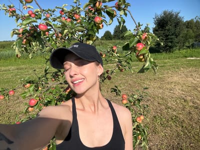 A woman in a black baseball cap and tank top smiles with her eyes closed while standing in front of an apple tree laden with red apples. The setting is outdoors, with a blue sky and green grass visible. The background includes trees and a field. The image appears to be a selfie taken on a sunny day. The location is Turovo, Russia.