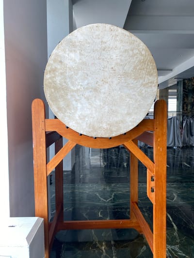 The image shows a large, round drum with a light-colored skin head, resting on a wooden stand. The stand is made of natural wood and has a simple, sturdy design with angled supports. The drum is positioned centrally in the frame.

In the background, the environment appears to be indoors, possibly a hall or a large room. The floor is dark and reflective, with a marbled pattern. There are some grey curtains hanging in the distance, and architectural elements like columns and lighting fixtures are visible. The walls are a light color.

There are no people visible in the image, nor are there any overt actions taking place. The scene is static and focused on the drum and its stand. The lighting suggests it is daytime, with light coming from the left side of the frame. The overall impression is one of quietness and potential use for cultural or religious purposes, given the nature of the object. The location is stated to be in the city of Aluekambuk, Indonesia.