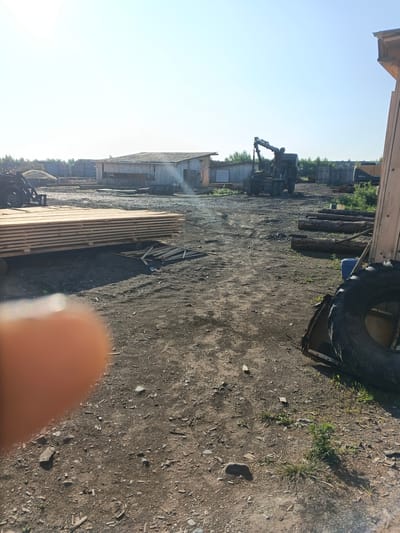 An outdoor, daytime, medium shot captures a lumber yard in Strelka, Russia. In the foreground, a stack of lumber rests on pallets. To the left, a person's thumb is blurred by its close proximity to the camera. Further back, a logging machine with a large arm is parked on the dirt ground. Several wooden buildings and a fence line the yard. In the background, trees and vegetation are visible. The sky is clear and blue.