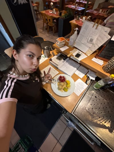 A young woman is posing for a photograph with a plate of food in a restaurant. She is wearing a dark-colored Adidas shirt with white stripes and a pink collar. Her hair is brown and pulled back. She is holding up two fingers in a peace sign.
On the table in front of her is a white plate with what appears to be Swedish meatballs, mashed potatoes with gravy, and lingonberry sauce. The table is covered with papers, a calculator, and other restaurant-related items.
The background shows the interior of a restaurant with tables and chairs. The restaurant could be in Bruchköbel, Germany.