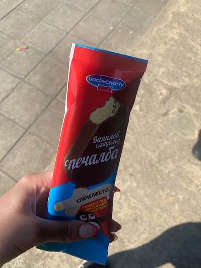 A human hand, likely female, with fingernails tipped in light pink polish, holds a red and blue ice cream wrapper. The wrapper features "Uncle Charly" in English, and Bulgarian Cyrillic text including "Ванилов сладолед" (Vanilla ice cream) and "спечаЛБА" (Win/Gain), with a stylized 'ЛБА' resembling 'ЧАРЛИ'. An image of a bitten chocolate-coated vanilla ice cream bar is prominent on the wrapper. Below this, further Bulgarian text reads "СПЕЧЕЛИХТЕ!" (You won!) and "ПРОВЕРИ СЛЕД КОНСУМАЦИЯ КЛЕЧКАТА!" (Check the stick after consumption!), indicating a prize promotion, alongside a small cartoon penguin. A timestamp "14:37:00" and "2023" are faintly visible on the wrapper. The background consists of light-colored, square-patterned concrete paving, illuminated by direct sunlight which casts a distinct shadow of a person to the right. A small, blurry, multicolored object (red and yellow/green) lies on the pavement in the upper left background. The scene is located in the city of Kipilovo, Bulgaria.