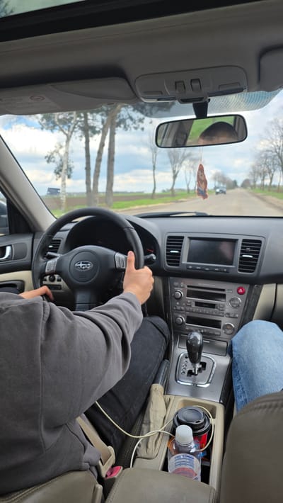 The scene depicts the interior of a Subaru vehicle, specifically the driver's seat perspective. A person with fair skin is driving, holding the steering wheel with both hands. They wear a gray hoodie and black jeans. A passenger wearing blue jeans is in the adjacent seat. The dashboard is visible, featuring a radio/entertainment system and gear shift. A cup of coffee and water bottles are in the central console. Outside the windshield and through the rearview mirror, a road lined with trees is visible. The scene is identified as taking place on a road in Senokos, Bulgaria, based on information provided.