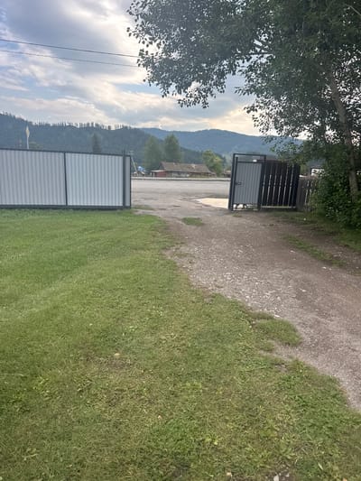 The image captures an outdoor scene near Yabogan, Russia, under a cloudy sky. In the foreground, a well-maintained lawn occupies the left side of the frame, with a dirt and gravel pathway to the right. Two gates are visible: a tall, corrugated metal gate on the left, and a partially open wooden gate on the right, revealing a darker, more solid gate behind it. Beyond the gates, a road is present, with a large puddle of water reflecting the sky. In the middle ground, several rustic wooden buildings with red roofs are situated. The background is dominated by rolling hills covered in dense evergreen trees. A large deciduous tree with lush green foliage is prominent in the upper right corner. Power lines are visible stretching across the sky. There are no people or animals present in the image.