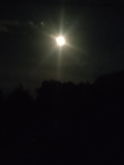 This image depicts a nighttime scene in Ternovka, Moldova, under a bright moon that is partially obscured by clouds. The moon's intense light creates a lens flare effect, radiating outwards. In the lower half of the frame, the dark silhouettes of trees and possibly buildings are visible against the faintly lit sky. The overall atmosphere is dark and somewhat mysterious, with the moonlight casting an ethereal glow. There is a small, bright point of light visible above and to the left of the moon, which could be a star or another celestial body. The scene appears to be a quiet, natural setting, with no people or specific objects clearly identifiable due to the low light conditions.