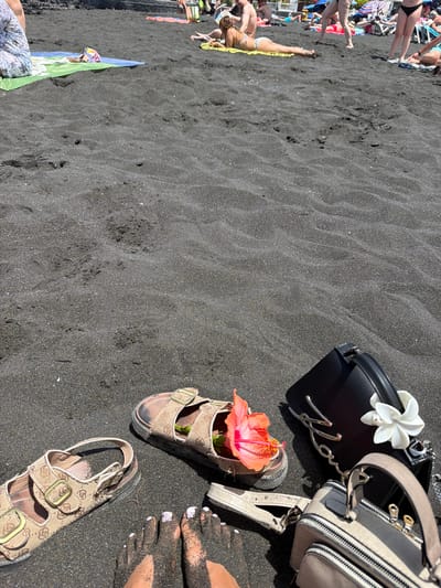 In Puerto de Santiago, Spain, on a black sand beach under a bright sky, a close-up shot focuses on a collection of items scattered on the dark, rippled sand. Two pairs of tan sandals, one with a prominent logo pattern and gold buckles, and another slightly more worn pair, sit near the foreground. A vibrant orange hibiscus flower is placed inside one of the sandals. To the right, two handbags are visible: a sleek black clutch with a silver logo chain and a decorative white floral embellishment, and a smaller, two-toned beige and gray crossbody bag with zippers and straps. In the bottom left corner, a person's feet are visible, with toes painted in light purple polish, partially covered in the black sand. In the background, other beachgoers can be seen lounging on towels and sunbeds, with umbrellas and a building complex suggesting a developed beach area.
