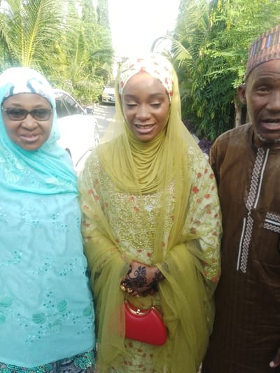 The image displays three individuals outdoors in Unguwa Uku Kauyen Alu, Nigeria. From left to right, an older woman, a younger woman, and an older man are positioned facing the camera. The older woman on the left wears black-framed spectacles and a light blue patterned hijab and garment, smiling slightly. The younger woman in the center wears an olive-green hijab with an underlying patterned headscarf, a sheer olive-green veil, and a matching olive-green dress featuring gold embroidery and red floral patterns. Her left hand exhibits intricate dark brown henna designs, multiple gold rings, and holds a small, red, structured handbag with a gold-colored clasp. She smiles with her eyes slightly downcast. The older man on the right wears a brown traditional garment with white and black embroidered trim and a multicolored patterned cap. His mouth is slightly open. The background consists of dense green foliage, including palm trees, and a partially visible white car on what appears to be a paved surface. The scene is illuminated by bright daylight.