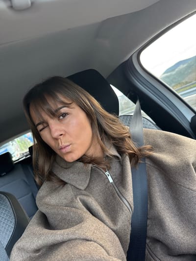 A woman wearing a brown coat and a seatbelt is photographed inside a car. She is looking toward the camera with a slightly puckered expression. The interior of the car is gray, with a view of the outside visible through the window. The photograph is taken at a slight angle. Her brown hair is partially obscuring one eye.