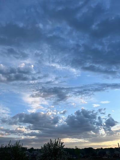 The image shows a sky filled with clouds, ranging from dark, ominous storm clouds on the left to lighter, more scattered clouds towards the right. The sky is predominantly blue, with patches of clear sky visible between the clouds. The photo seems to have been taken near Зыково, Russia, as evidenced by the presence of trees and rooftops along the top and right edges, typical of a populated area. The lighting suggests either early morning or late evening. There are no people or subjects immediately visible within the frame.