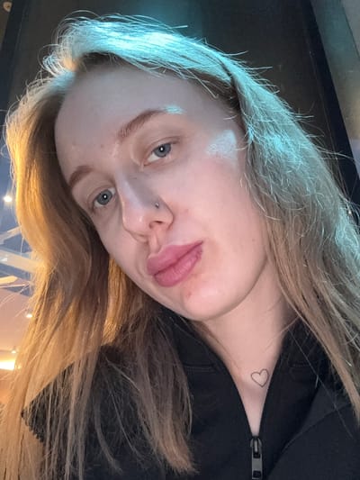 A close-up, rotated portrait of a fair-skinned woman with blonde and brown hair and blue eyes. She has a nose piercing. A small heart tattoo is visible on her neck, near the zipper of a black garment she is wearing. The environment is indistinct but appears to be indoors, with some ambient lighting. There is no indication of Ol'ginskaya, Russia in this image.
