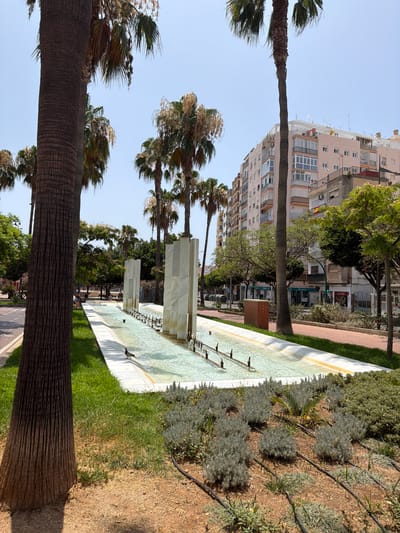 The image depicts a long, shallow water fountain situated in a public space, likely a plaza or park, in Almería, Spain. The fountain features a long, rectangular basin with clear water flowing and multiple small spouts creating gentle jets. The basin is lined with white marble-like material and a yellow trim. Several large, smooth, white rectangular structures rise from the center of the fountain. Palm trees with rough, textured trunks and fronds are prominent throughout the scene, framing the fountain and lining the background.

In the background, a multi-story building with a light pink facade and numerous balconies dominates the skyline. Other buildings of varying heights and architectural styles are visible, suggesting an urban environment. The street runs alongside the fountain, with a road and sidewalk visible. Green grass covers the area immediately surrounding the fountain, and a bed of low-lying grey-green plants, possibly lavender, is located in the foreground, with visible irrigation lines. A single pigeon is standing on the edge of the fountain's white lining. The sky is clear and blue, indicating a sunny day. The overall impression is a well-maintained urban public space with a focus on greenery and water features.