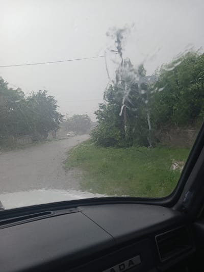 The image, taken from inside a vehicle, depicts a view through a rain-streaked windshield. Outside, a dirt road winds through a rural area characterized by lush green trees and vegetation. The sky is overcast and grey, indicating rainy weather. In the distance, a white car is visible on the road. The foreground shows the interior of the car, including the dashboard. The scene appears to be a snapshot of a journey on a road in Skrebyshi, Ukraine, during a downpour.