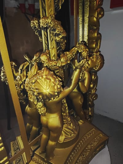 This is a medium shot, eye-level, horizontal photograph of a golden, decorative statue with cherubic figures. The statue is ornate and appears to be made of a metallic material, possibly resin or painted metal, with a matte gold finish. Several cherubic figures, depicted as plump infants with curly hair, are intertwined with floral elements, specifically roses. One cherub is shown with its head tilted back, reaching up towards a rose, while another is positioned behind it, partially obscured. The base of the statue is also detailed with decorative carvings and appears to be part of a larger structure. The lighting creates highlights and shadows, emphasizing the texture and sculpted details of the figures and ornamentation. The background is indistinct and appears to be an indoor setting with a wall on the right and darker, shadowed areas to the left, including what might be furniture or another object. There are no people present in the photograph besides the sculpted figures. The image does not contain information that can determine its location in the city of Kirrwiller, France.