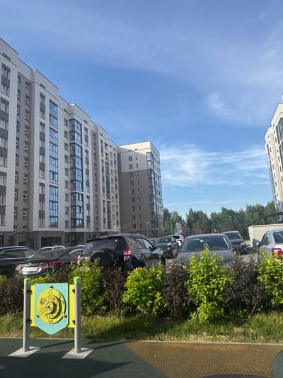 The image shows a modern residential complex with multi-story buildings. There are several cars parked in front of the buildings, including SUVs and sedans. Lush green bushes and trees surround the area. In the foreground, there is a children's playground equipment with colorful panels and posts. The sky is blue with scattered clouds. The scene appears to be in Koltsovo, Russia, given the context. There are no people visible in the image.