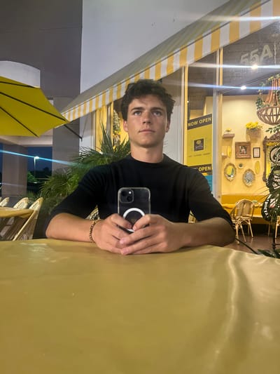 A young adult male, with dark, wavy hair, is seated at an outdoor patio table during nighttime in front of Joelle's Deli Cafe, located at 55A Peninsula Center, Rolling Hills Estates, California, United States. He is wearing a black long-sleeved shirt and gazes slightly upwards and to his right. His hands are clasped on the golden-yellow tablecloth, holding a smartphone with a clear case and a visible MagSafe ring. A beaded bracelet is on his left wrist. To his left, empty wicker chairs with light cushions and a yellow patio umbrella are visible. The cafe's storefront features a yellow and white striped awning and large glass windows. Through the windows, the brightly lit interior displays yellow walls, decorative mirrors, a wooden chandelier-like fixture, potted plants, and matching wicker chairs, along with a "NOW OPEN" sign for "Joelle's Deli Cafe - Flowers - Bakery". Streetlights are visible in the distance beyond the patio area.