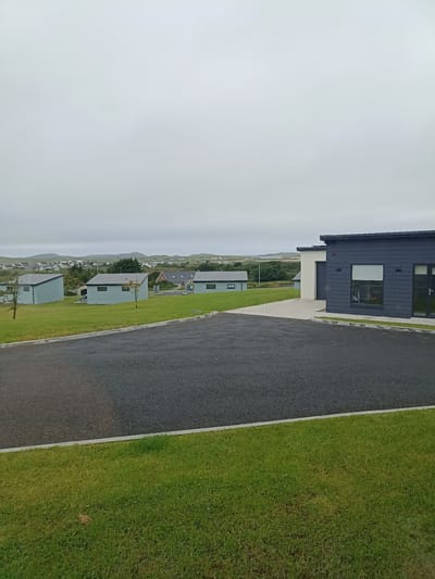 The image depicts a street scene in Doirí Beaga, Ireland, with a focus on a driveway and several modern, single-story structures. The foreground features a dark asphalt driveway bordered by a strip of green grass. Beyond the driveway, a paved area leads to the entrance of a dark-colored modern building with large windows and a garage door. To the left of this building, several light blue, rectangular structures with gray roofs are visible on a grassy slope, interspersed with small trees. In the background, rolling hills and a scattering of houses indicate a semi-rural or suburban setting, with a hazy sky suggesting overcast weather. There are no people or vehicles visible in the frame.
