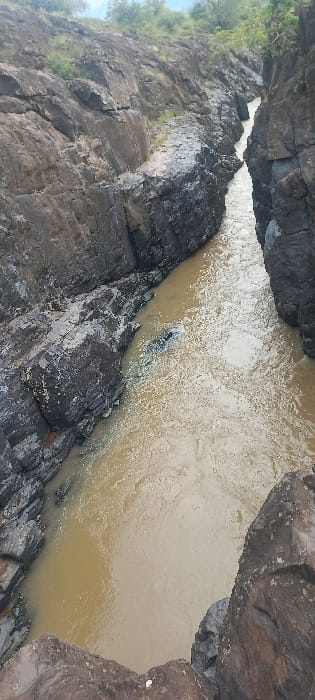 This image depicts a section of a river flowing through a narrow, steep-sided rock gorge in Ainamoj Village, Kenya. The river water is a murky brownish-tan, exhibiting visible surface ripples and patches of lighter foam, indicating current flow. The gorge walls are composed of dark, rugged rock formations, rising abruptly from the water on both sides. These rock surfaces display varied textures, from large, fractured blocks to smoother, weathered sections. Near the left bank of the river, approximately midway along the visible stretch, a dark, elongated object is partially submerged in the water, creating a slight disturbance on the surface. Sparse green vegetation is discernible along the upper rims of the canyon, with a hazy or overcast sky visible beyond. The photograph is taken from an elevated vantage point, looking down into the gorge and along the river's course. There are no individuals present.