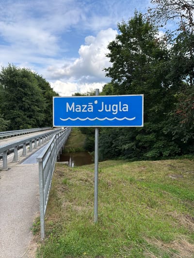 The image shows a blue sign with white text and wavy lines, indicating a location name. The sign reads "Mazā Jugla" which translates to "Little Jugla". The sign is positioned vertically and is attached to a metal pole. It is situated outdoors, next to a body of water and a grassy slope. A metal railing of a bridge or walkway runs along the foreground of the image, with a concrete path beside it. Lush green trees are visible in the background, and the sky is partly cloudy with some white clouds against a blue backdrop. The overall scene suggests a scenic outdoor environment, possibly a park or a recreational area near a river or canal. The lighting suggests it is daytime, and the weather appears to be pleasant, with good visibility. There are no people visible in the image.