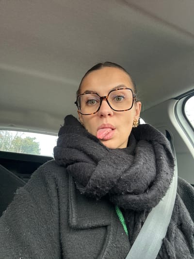 A female individual is depicted in a close-up selfie taken inside a vehicle in Saint-Cyprien, France. She has light skin, light green eyes, and light brown hair pulled back from her face. She wears large, square-framed tortoiseshell eyeglasses. Her tongue is extended from her mouth in a direct, forward pose. A small gold stud earring is visible in her left ear, and a larger, decorative gold earring is present in her right ear. She is dressed in a dark, textured coat, and a thick, dark scarf is wrapped around her neck. A strip of bright green fabric is visible beneath the scarf. A light gray seatbelt diagonally crosses her chest. The vehicle's interior features a light-colored headliner. Through the rear window, blurred green foliage and an overcast sky are visible.