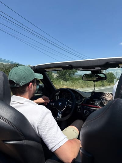The image shows a view from the back seat of a convertible car. The primary subject is a person wearing a green baseball cap and a white shirt, seated in the driver's seat. They appear to be reaching for something on the dashboard. Sunlight streams in, indicating it is daytime. The car's interior is visible, including the black leather seats, steering wheel with a BMW logo, and part of the dashboard. Outside the car, a clear blue sky is visible with some power lines in the foreground. The background shows a rural landscape with trees and hills under the bright sky, suggesting a sunny day in a potentially rural or semi-rural location. The overall mood is casual and relaxed, consistent with a scenic drive on a pleasant day. No visible text is present in the image.