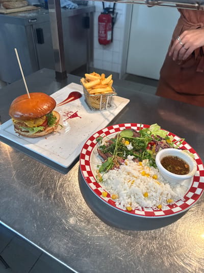 In a Rivesaltes, France restaurant, a close-up shot captures a meal served on a stainless steel countertop. The meal features a hamburger with a skewer, fries in a metal basket, and a plate of rice, salad, and sauce. A person's hands, adorned with rings, are visible in the upper left corner, suggesting they are about to partake in the meal or have just finished. The restaurant's interior, with white tiled walls and a fire extinguisher, is partially visible in the background. The scene appears to be set during daytime, and the overall mood is casual, hinting at a relaxed dining experience. There is no visible text in the image.