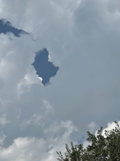 The image shows a sky with scattered clouds and a patch of blue sky in the center. There is a tree branch with green leaves in the upper right corner of the image. The sky appears overcast with some bright spots indicating sunlight. The overall impression is that of a partly cloudy day, possibly in the late afternoon given the diffused light. No people, objects, or text are visible in the image. The scene is a natural outdoor setting, looking up towards the sky.