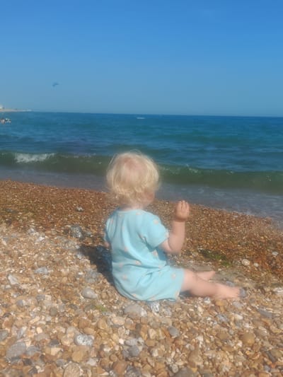 A young child with blonde hair, wearing a light blue dress with a pattern of small white shapes, sits on a pebble beach facing the ocean. The child is viewed from behind and is sitting with their legs extended. The beach is composed of small, rounded pebbles in various shades of brown, grey, and white. In the background, the turquoise ocean is calm with small waves breaking near the shore. The sky is a clear, bright blue with a faint kite visible in the distance. The scene is set on a sunny day. This location is likely Worthing, United Kingdom, given the type of beach and sea.