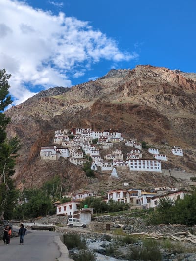 This is a medium-shot, vertical, eye-level, full-color photograph depicting a Tibetan Buddhist monastery nestled on the side of a rocky mountain in Yulang, India. The monastery's white, multi-storied buildings with dark-trimmed windows are spread across the mountainside, appearing as a collection of connected structures. The mountain itself is rugged and barren, with shades of brown and ochre dominating its surface, and sparse green vegetation clinging to its lower slopes. A clear blue sky with scattered white clouds forms the backdrop. In the foreground, a paved road curves through the scene, with three individuals, two adults and a child, walking on it. To the right of the road, a white SUV is parked near a cluster of buildings, and a rocky stream flows beside the road.