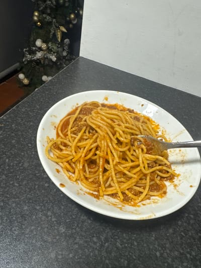 A partially eaten plate of spaghetti with meat sauce sits on a dark gray countertop. A silver fork is inserted into the noodles. Part of a Christmas tree decoration is visible in the lower left corner of the image. The countertop is next to a white surface. The location is Warrington, United Kingdom. There are no people visible in the image.
