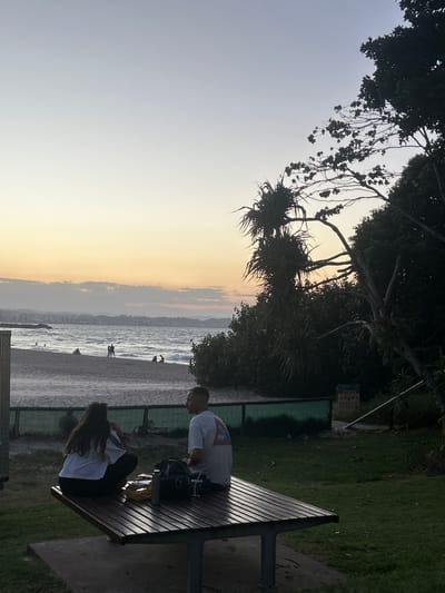 The image captures a serene coastal scene at dusk, likely in Coolangatta, Australia. The sky transitions from a soft blue-grey at the top to warm golden and orange hues closer to the horizon, indicating a late afternoon or sunset. Overhead, dark, silhouetted trees, some resembling pandanus with spiky leaves, frame the upper portion of the view, their branches reaching into the clear sky.
In the mid-ground, a wide stretch of ocean displays gentle waves, with a few distant figures visible in the water and on the sandy beach. A hazy landmass or cityscape is discernible across the water in the far distance.
To the right, in the foreground, two individuals are seated at a wooden picnic table. A man, wearing a light-colored t-shirt, sits facing the ocean, seemingly engaged with a dark bag or item on the table. Beside him, a person with long dark hair, wearing a white t-shirt and dark pants, is also seated, with their back mostly to the camera, looking down at the table. The slats of the picnic table show distinct light and shadow patterns, illuminated by the low sun.
Behind the picnic table and to the left of the grassy area, a notice board is visible. Text on the sign indicates "COOLANGATTA" at the top, along with various prohibition pictograms (such as no dogs, no littering). A larger blue section below has text "CITY OF GOLD COAST" and a phone number that appears to be "1300 GOLDCOAST" (1300 465 326). The overall atmosphere is peaceful and relaxed.