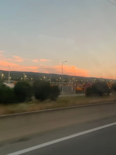 A slightly blurry, wide-angle, eye-level shot captures a rural scene during sunset in Gorovani, Georgia. The sky is dominated by a gradient of soft blues and oranges, with streaks of pink clouds scattered across the horizon. In the mid-ground, a line of hills creates a silhouette against the colorful sky. Below the hills, a series of tall streetlights with yellow bulbs illuminates what appears to be a construction site or industrial area. Yellow construction vehicles are visible among the structures. In the foreground, dense green foliage forms a dark, blurry band across the lower third of the image. A paved surface, possibly a road or highway with a white line, runs horizontally along the bottom of the frame, also appearing slightly blurred due to motion. The overall impression is of a vast, open landscape at dusk.