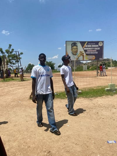 Two young men are captured in an outdoor, daytime scene, likely in Yuli Campus, Nigeria, with a bright blue sky and scattered clouds. One man, on the left, stands and looks towards the camera, wearing a white t-shirt and jeans. The other man, on the right, is also in jeans and a white t-shirt, with headphones visible. He is leaning slightly and looking down. Behind them, a large billboard welcomes visitors to "SA'ADU ZUNGUR UNIVERSITY (SAZU) YULI CAMPUS" and features an image of Sen. Bala Abdulkadir Mohammed CON, the Executive Governor. The ground is a dirt surface, with some grass visible to the left. In the background, a sports field with a net and a few other people can be seen. The overall atmosphere appears casual and relaxed.