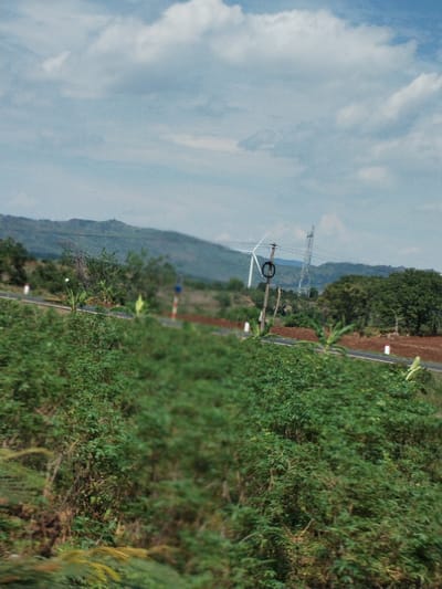 The image depicts a rural scene in Thôn 4, Vietnam. In the foreground is dense green vegetation, possibly shrubs or bushes, blurring the view. A road cuts through the middle ground, marked with white posts. Beyond the road, the landscape rises to rolling hills covered in sparse vegetation. A wind turbine and an electrical transmission tower are visible in the distance, suggesting a source of power generation. The sky is partly cloudy with a light blue hue.