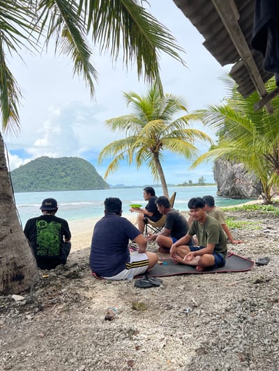 A group of young men are gathered on a rocky beach in Kruengtunong, Indonesia. The scene is tropical, with palm trees framing the shot and a lush, green hill rising from the turquoise ocean in the background. The sky is partly cloudy, suggesting it is daytime.

Five men are visible, most of them sitting on the ground or a black mat. One man in a black long-sleeved shirt with a green graphic design and a black baseball cap stands slightly apart, looking out towards the water. Another man, wearing a navy blue t-shirt and white shorts with yellow stripes, is also looking towards the ocean, holding a green frisbee. The other men are seated closer together, some appearing to be conversing or observing. One man in a green t-shirt is holding what appears to be a cigarette.

The overall mood seems relaxed and casual, consistent with a beach gathering. The presence of palm trees, the clear water, and the tropical vegetation strongly indicate a warm, possibly humid climate. The terrain is composed of small rocks and pebbles, typical of many Indonesian coastlines. No specific event is clearly depicted, but the presence of the frisbee suggests recreational activity. The lighting suggests it is likely late morning or early afternoon.

There is visible text on the black long-sleeved shirt of the standing man, featuring a complex green graphic. The baseball cap also has some text, possibly "typen".