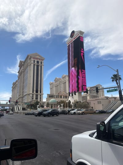 The image captures an outdoor scene in Paradise, Nevada, featuring Caesar's Palace with a digital billboard promoting Nike, visible against a blue sky with scattered clouds. Traffic lights and palm trees line the street, and parked cars are adjacent to the sidewalk. In the foreground, a white vehicle with a side mirror is partially visible, suggesting the viewpoint is from inside a car. The overall impression is a daytime view of the Las Vegas Strip, combining commercial advertising with urban architecture.