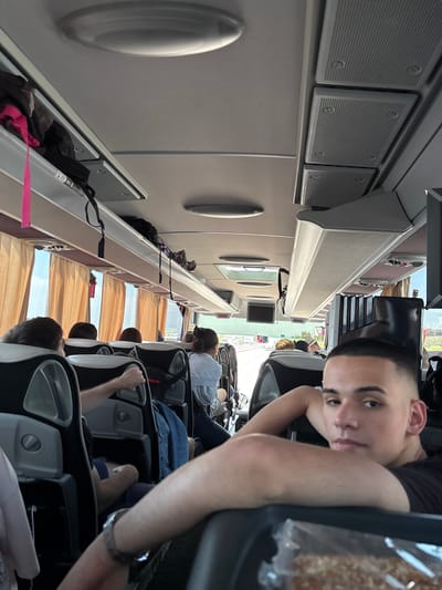 The image shows a man with a short haircut on a bus. He is seated and looking at the camera. The bus appears to be in transit, judging by the other passengers and the scenery visible through the windows. The interior of the bus has overhead storage compartments and individual seats.
The bus is in Noventa di Piave, Italy. This is determined from the view through the window.