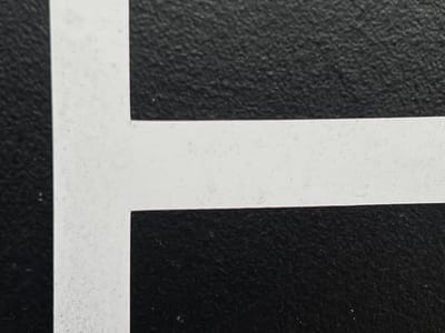 The image is a close-up, abstract shot of a white line on a textured black surface. The white line appears to be painted or adhered to the surface. It has a slightly rough texture with some small imperfections. The black surface has a noticeable grain or pattern. The image is a detail of a larger composition, likely a boundary or marking. There are no people, animals, or identifiable objects present. The lighting appears to be even, with no strong shadows. The location in Lanžhot, Czechia, is not discernible from the image itself.