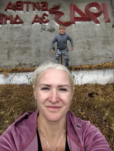 In Balyko-Shchuchynka, Ukraine, a woman takes a selfie in the foreground, smiling. She has blonde hair and is wearing a purple hooded jacket. Behind her, a young boy in a grey hoodie and camouflage pants stands with his arms outstretched, appearing to jump or pose. They are positioned in front of a weathered concrete wall adorned with large, red, three-dimensional letters that spell out "ЧОРНОБИЛЬ" (Chernobyl). The ground in front of the wall is covered with dry grass, fallen leaves, and some thin branches. The environment suggests an outdoor, possibly historical or memorial, location.