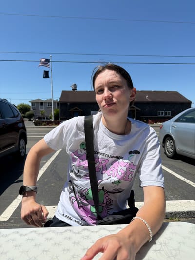 The image captures a person sitting at an outdoor table, likely at a restaurant or cafe, on a sunny day in Cannon Beach, Oregon, United States. The background reveals a street scene with parked cars, buildings, and a flagpole flying the American flag and another flag below it. The person, who appears to be a young woman, is wearing a white t-shirt with a graphic print and a black crossbody bag. She has a watch on her left wrist and a bracelet on her right wrist. Her expression is directed towards the camera. The foreground features the edge of a white patterned table. The overall environment suggests a casual outdoor setting with a clear blue sky overhead.