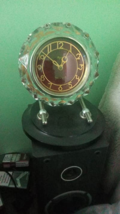 The image shows a vintage clock with a burgundy face and gold numbers and hands, mounted on a black base. The clock has a decorative crystal outer casing. The clock is positioned on top of a black object, which appears to be a speaker. To the left of the speaker, a pack of cigarettes is visible. The background is a plain green wall. The scene is indoors and dimly lit. There is no indication of the city of Pereiaslav, Ukraine in the image.
