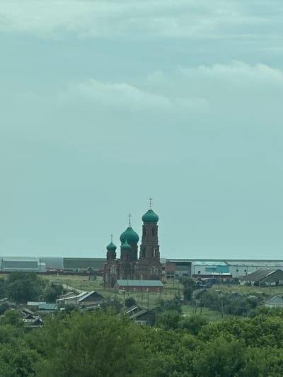 An elevated, medium-shot captures a view of a village in Yekaterinovka, Russia, with a prominent brick church with green domes in the background. The sky is overcast and a pale blue, dominating the upper half of the frame. Below the sky, a hazy line separates the sky from the landscape. In the mid-ground, the church stands out with its brick facade and three distinct green-domed towers, each topped with a cross. To the left and right of the church, several low-rise buildings with grey and blue roofs are visible, suggesting an industrial or agricultural area. The foreground is filled with lush green trees, obscuring the lower portion of the village buildings. The overall impression is one of a rural settlement under a cloudy sky, with the church as a central architectural element. There are no people or animals visible in the scene.