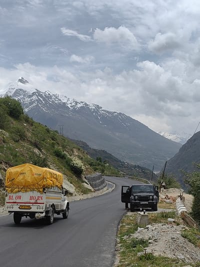 In a mountainous region of Unknown, India, an overcast sky looms over a winding asphalt road. The road curves through a valley flanked by steep, vegetation-covered slopes leading to snow-capped mountains in the distance. A white pickup truck with a yellow tarp-covered load, bearing the words "BL OW HORN" and license plate "HP 69 7756," travels along the road. Further ahead, a dark SUV with its doors open is parked on the right side of the road, with a person standing beside it. The environment is characterized by rugged terrain, rocky outcrops, and scattered greenery.