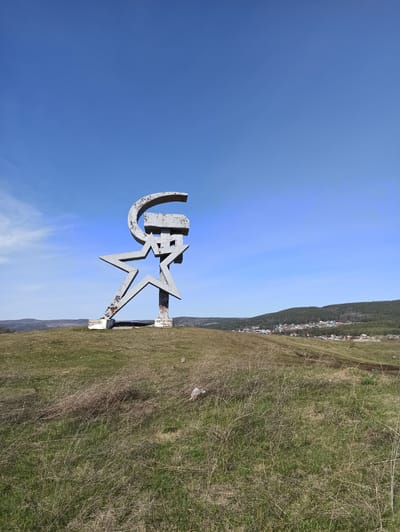 The image shows a monument featuring a star and the letters "GR" (presumably standing for "Gorno-Rudnaya Kompaniya" or Mining and Metallurgical Company) set against a clear blue sky. The monument is made of weathered metal, standing on a grassy hillside overlooking the city of Satka, Russia. In the background, a dense residential area can be seen at the foot of forested hills. The photograph is taken from a low angle, emphasizing the size and prominence of the monument within the landscape.