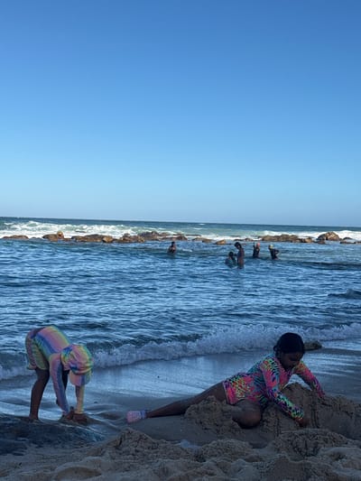 Two children are playing on a beach in Muizenberg, South Africa. One child, wearing a colorful patterned rash guard and pink shoes, is climbing a sand dune on the right side of the frame. Another child, wearing a rainbow-striped rash guard, is crouched near the water's edge, also on the right side. In the background, a line of rocks is visible along the coastline, with several people standing in the water near the rocks. The ocean waves are crashing against the rocks and the shore. The sky is clear and blue on the left side of the frame.