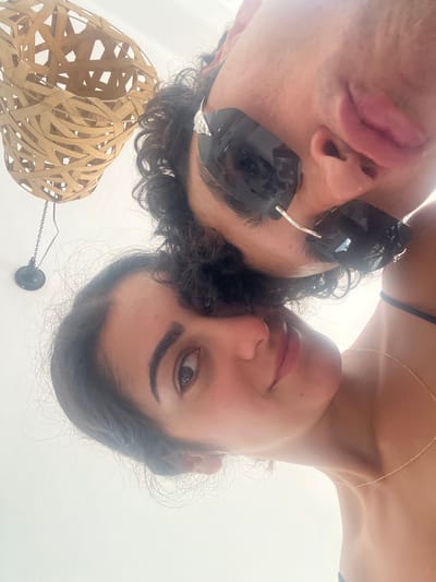 The image is a close-up selfie taken from a low angle, capturing two individuals, a man and a woman, in an intimate pose. The man, in the foreground and slightly above the woman, is wearing black eye patches and dark sunglasses. His hair is curly and dark. His lips are slightly parted. The woman is below him, her face is closer to the camera. She has dark hair and her eyes are open and looking directly at the camera. She is wearing a thin gold necklace. In the background, a portion of a woven light fixture made of natural materials is visible, hanging from a chain. A small black spherical object is attached to the chain. The overall environment appears to be indoors with bright, diffused lighting, suggesting a daytime setting. The image lacks any specific visual cues that would identify it as being located in Ceglie Messapica, Italy.