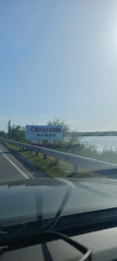 The image, taken from inside a vehicle, shows a sign indicating the "Salsky District" in Russian. The sign is white with red lettering. The car is driving on a road adjacent to a body of water, likely a river or lake, with greenery on the shoreline. The sky is blue and clear. The scene appears to be located in a rural or semi-rural area, likely near the city of Манычстрой, Russia, based on the location provided.