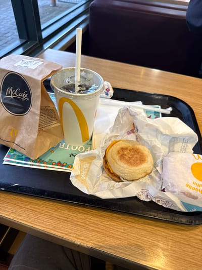 The image depicts a meal served on a black plastic tray inside a McDonald's restaurant. The exact location within East Hampshire, United Kingdom, cannot be determined from the visual information provided. On the left side of the tray is a brown McCafé branded paper bag, with a white label indicating "Sugar Donut." Central to the tray is a clear plastic cup containing a dark carbonated beverage with ice and a white paper straw, displaying the McDonald's arch logo. To the right, an unwrapped breakfast sandwich, consistent with an Egg McMuffin (comprising an English muffin, a circular patty, and melted cheese), rests within its white paper wrapper. A second, partially wrapped item, also appearing to be a breakfast muffin, is visible to its right. Underneath the paper bag and drink cup, a green promotional sheet with the partially visible word "FOOTB" is present. The black tray rests on a light-colored wood-grain table. In the lower left foreground, the leg of a person wearing dark trousers is visible beneath the table. The background features a large window showing a blurred exterior and a dark red upholstered booth seat, indicating an indoor dining environment during daylight hours.